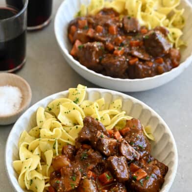 Two bowls containing beef bourguignon and buttered noodles with freshly chopped parsley.