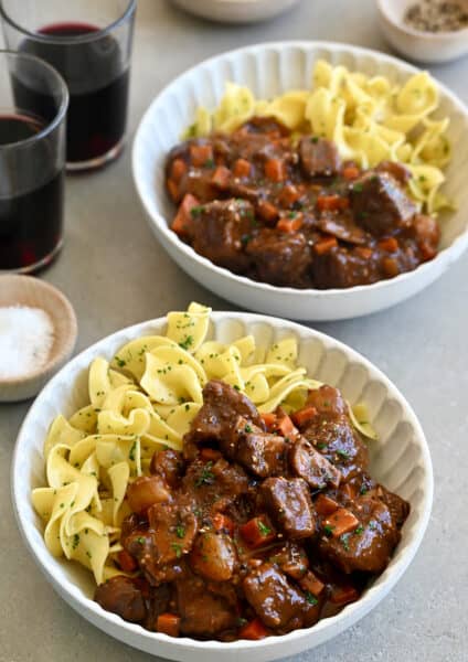 Two bowls containing beef bourguignon and buttered noodles with freshly chopped parsley.