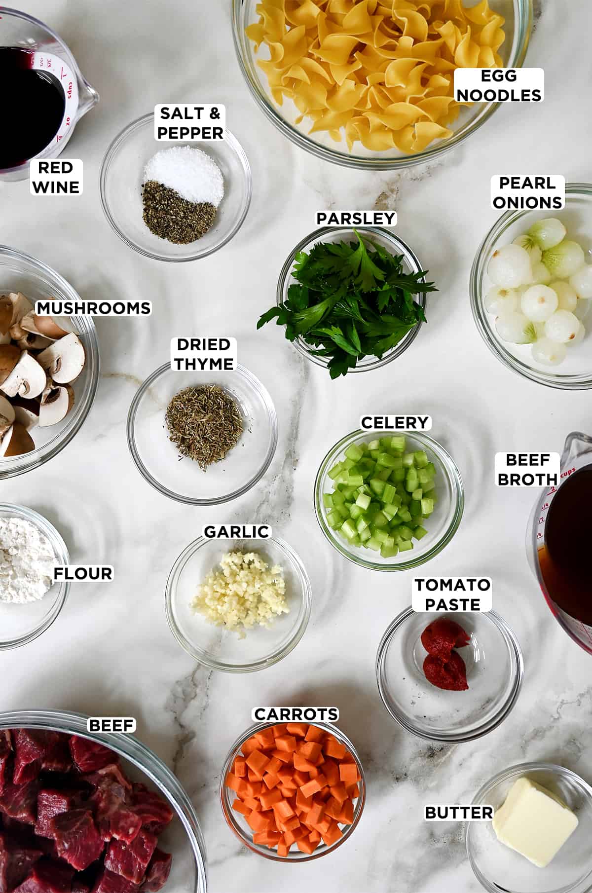 Various sizes of clear bowls containing red wine, egg noodles, fresh parsley, butter, pearl onions, diced celery, dried thyme, mushrooms, cubed beef, salt and pepper, tomato paste, diced carrots, beef broth and minced garlic.