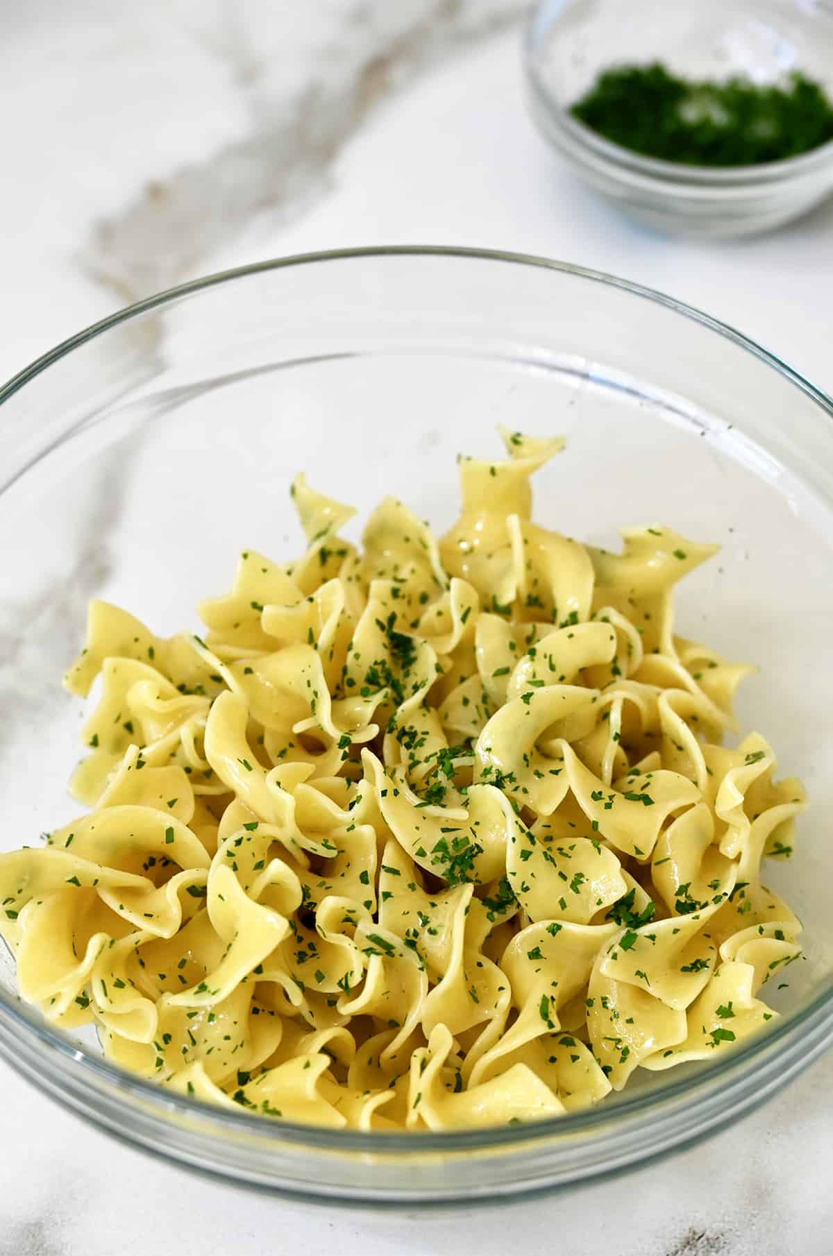 A clear bowl containing buttered noodles garnished with chopped fresh parsley.