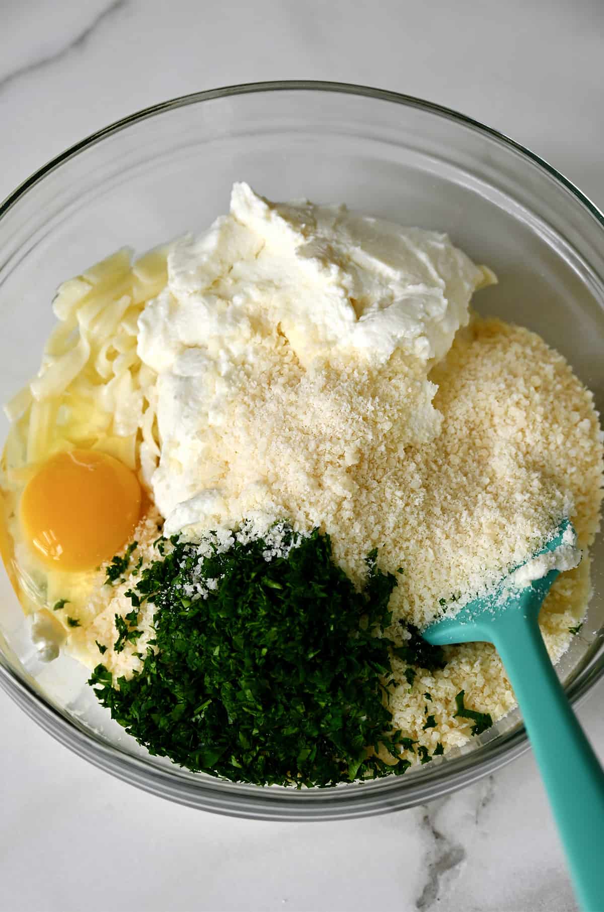 A glass bowl containing ricotta cheese, shredded mozzarella cheese, grated Parmesan cheese, minced fresh parsley and an egg.