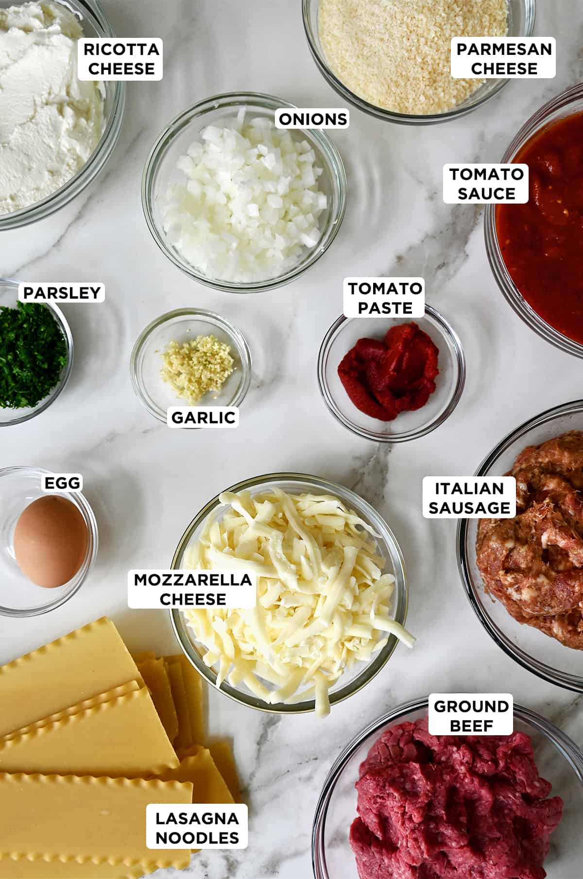 Various sizes of clear bowls containing all of the ingredients needed to make lasagna, including ricotta cheese, diced onion, Parmesan cheese, tomato sauce, tomato paste, Italian sausage, ground beef, mozzarella cheese, lasagna noodles, an egg and fresh parsley.