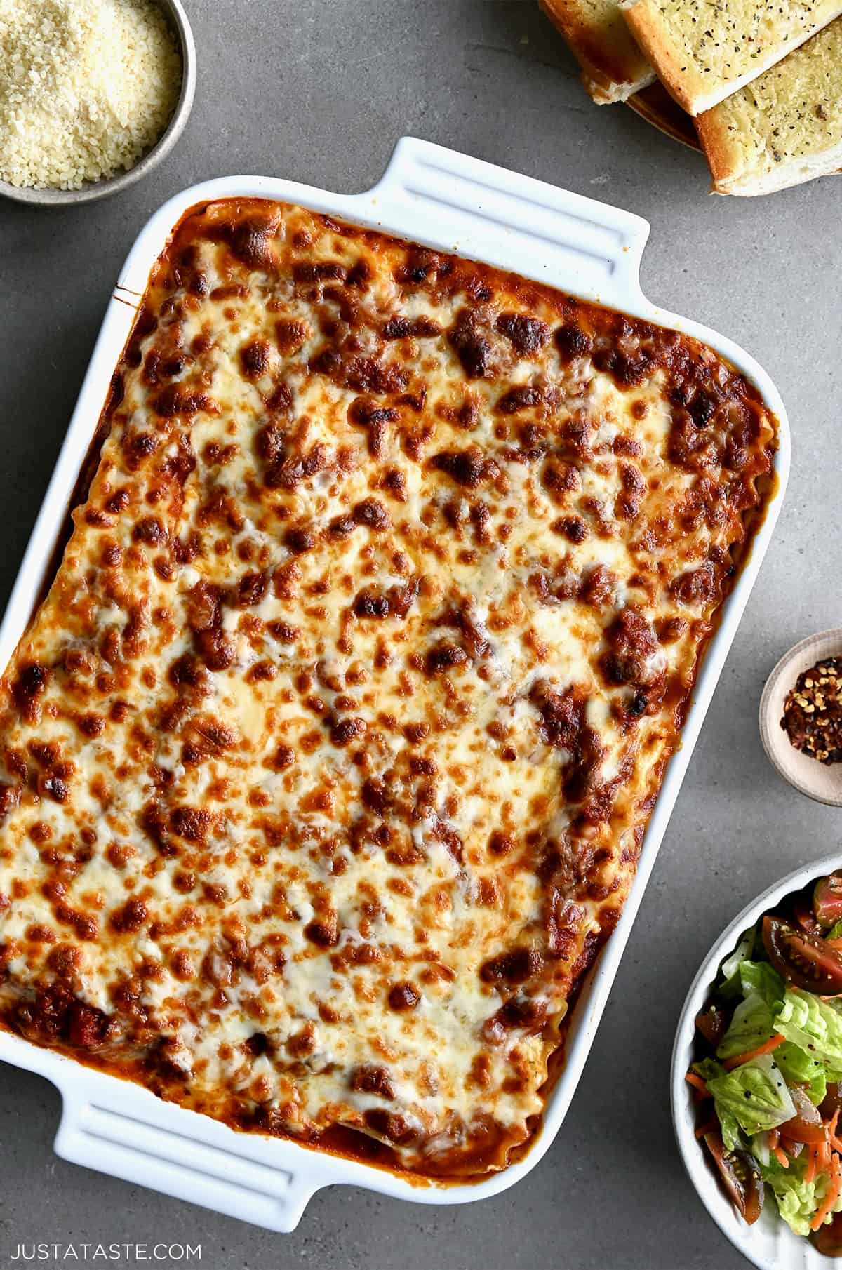 A white 9x13 baking dish containing homemade lasagna with a golden cheese crust next to a bowl containing a crisp green salad.