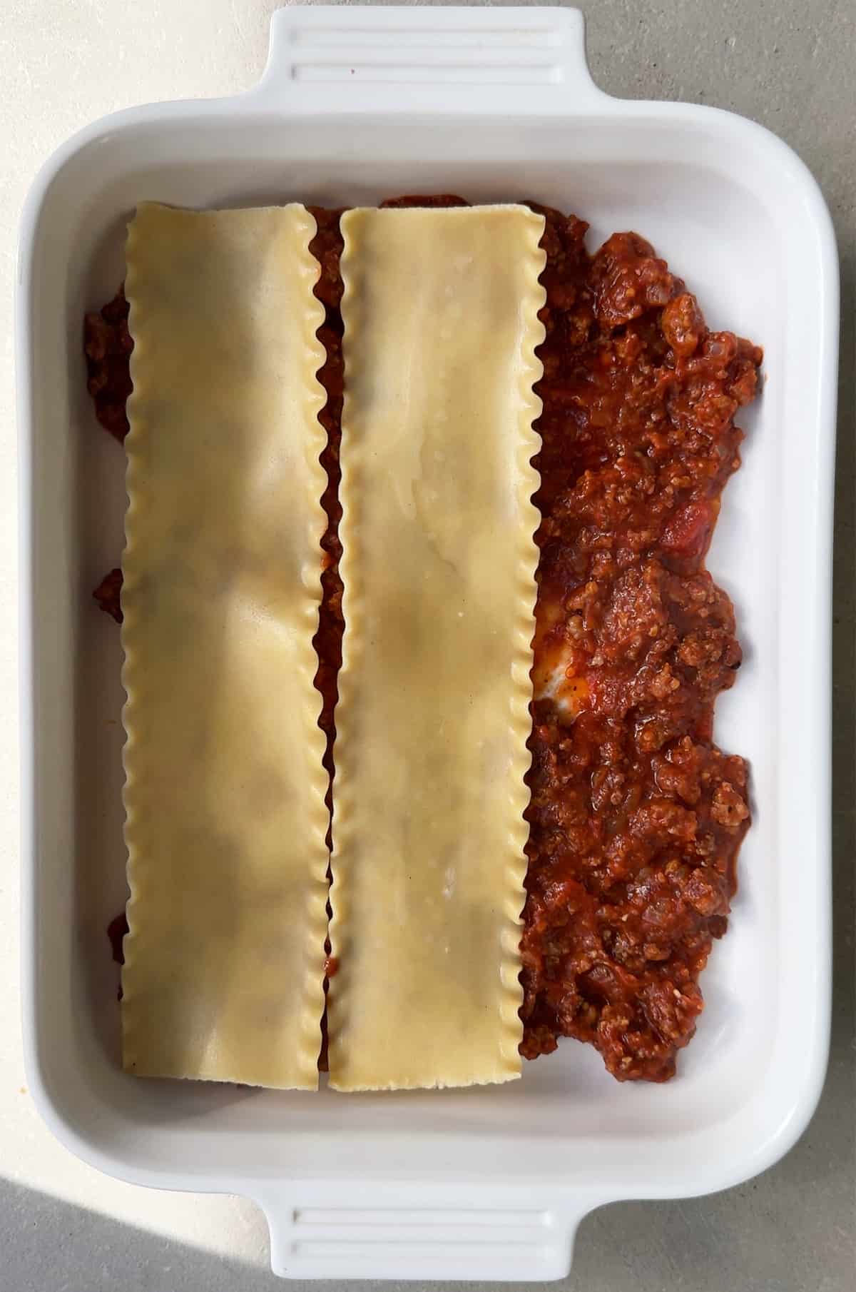 A white baking dish with meat sauce topped with two lasagna noodles.