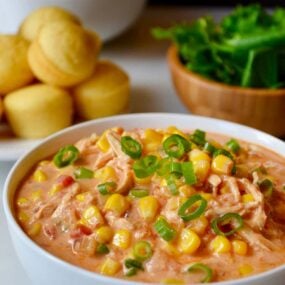 A white bowl containing Salsa Corn Chowder with a plate of corn muffins behind it