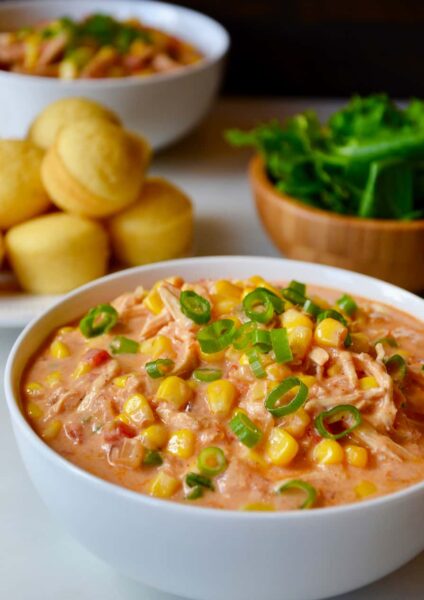 A white bowl containing Salsa Corn Chowder with a plate of corn muffins behind it