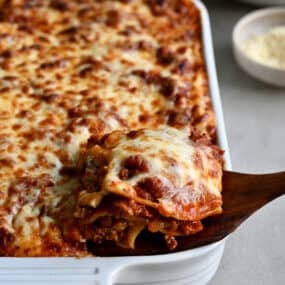 A wooden spatula lifts a slice of lasagna out of a baking dish.