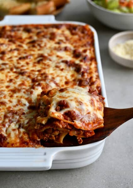 A wooden spatula lifts a slice of lasagna out of a baking dish.