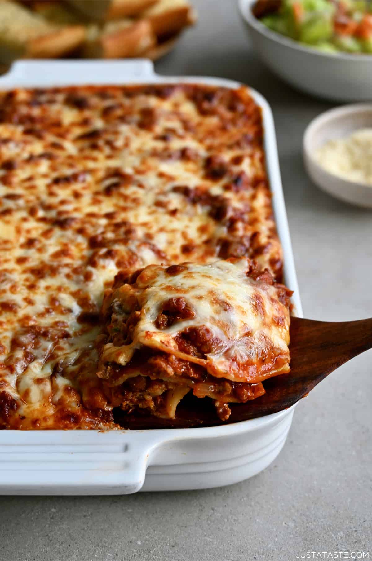 A wooden spatula lifts a slice of lasagna out of a baking dish.
