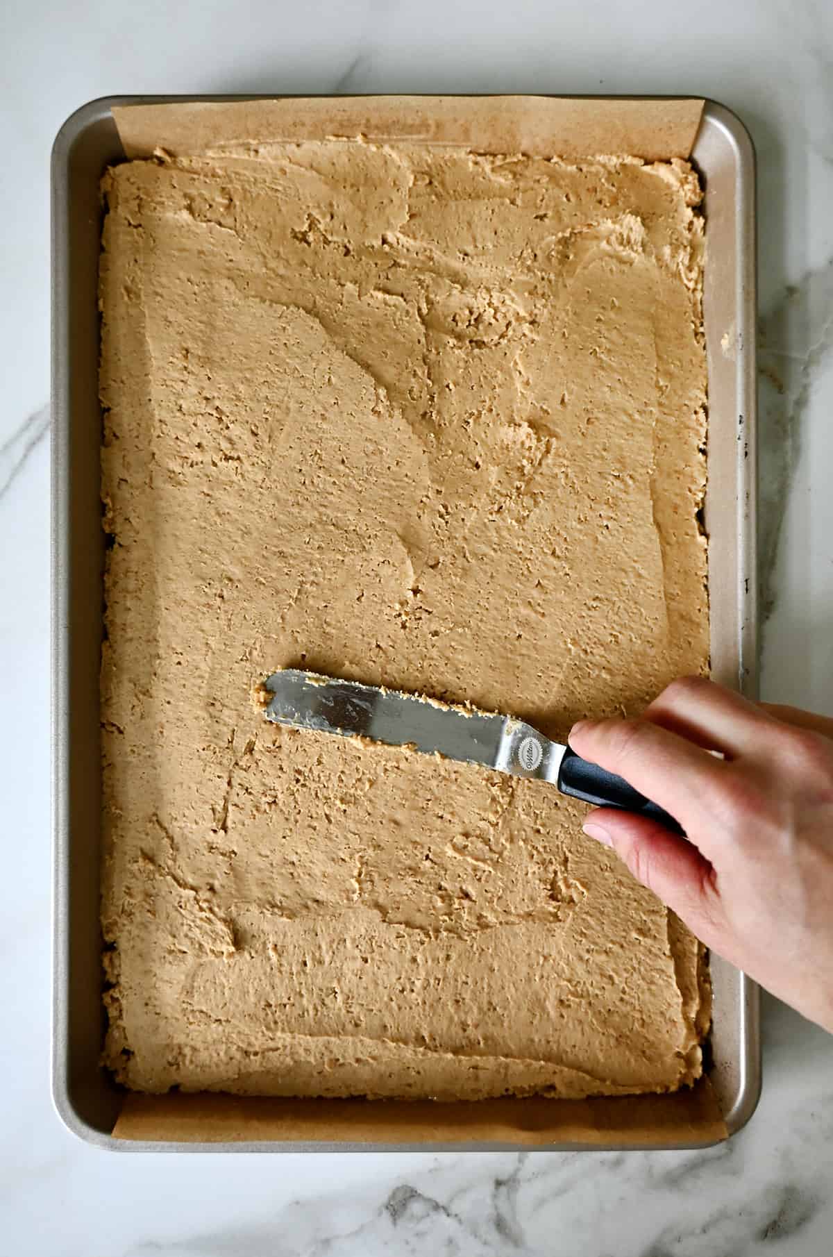 A hand holding an offset spatula spreads peanut butter bars into an even layer on a baking sheet.