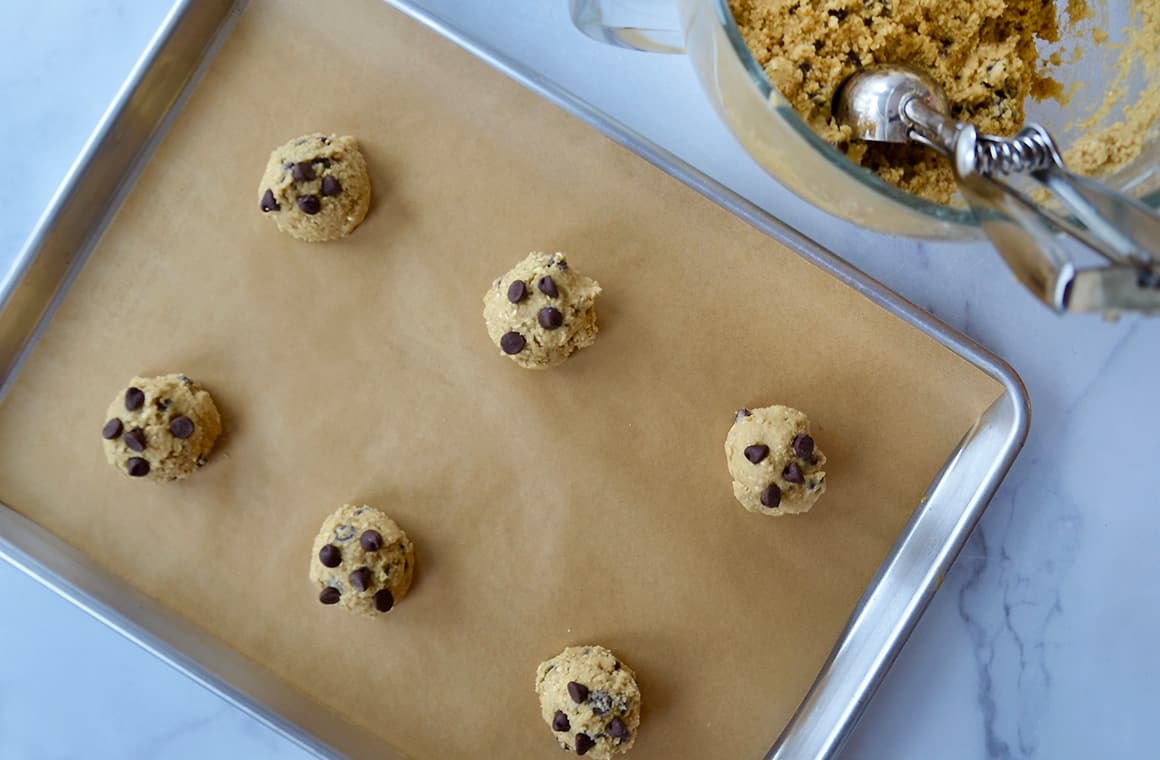 Scooped balls of cookie dough on parchment paper-lined baking sheet next to bowl containing cookie dough and scoop