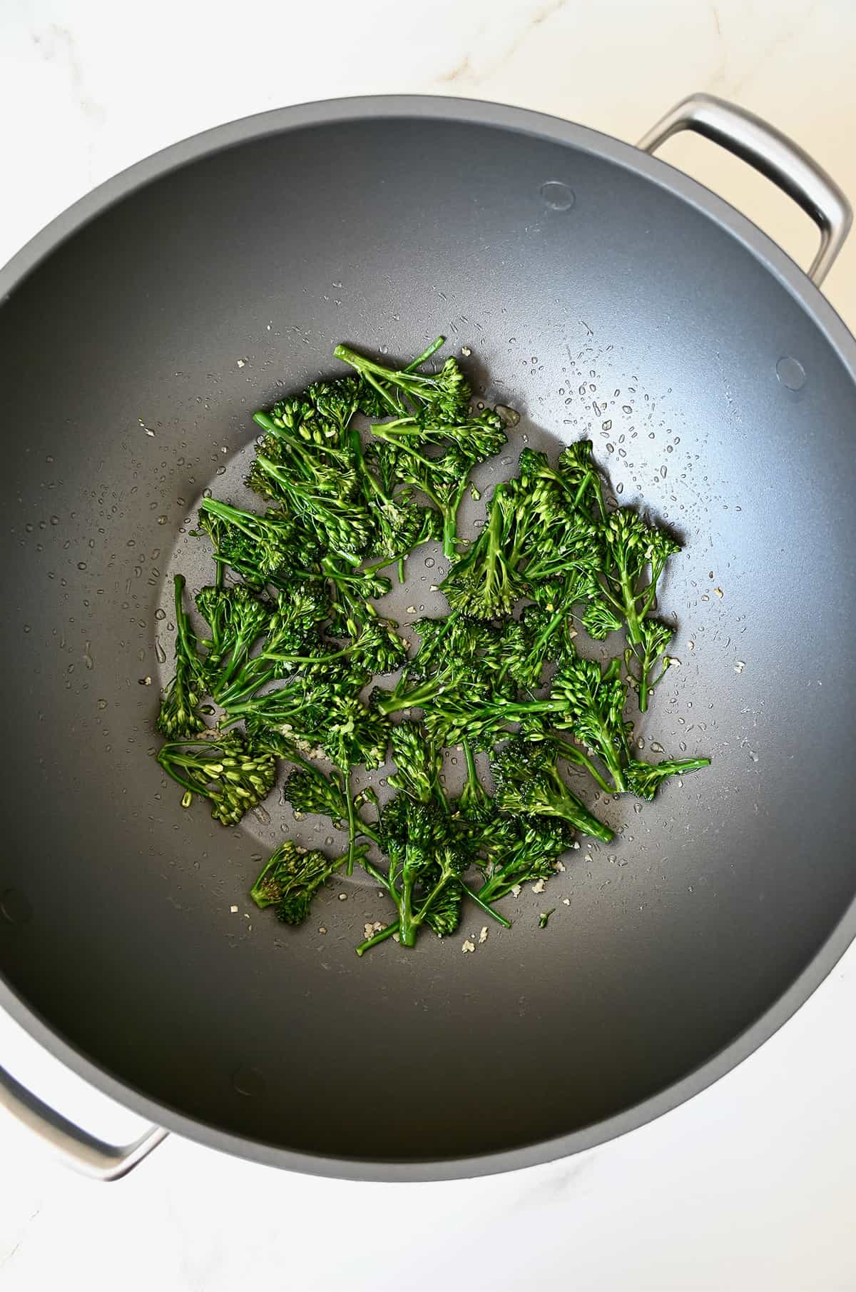 Broccolini pieces in a wok with minced garlic and oil.