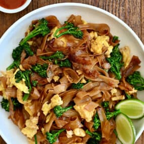 Pad See Ew with lime wedges on a white dinner plate next to a small bowl containing sweet and sour sauce.