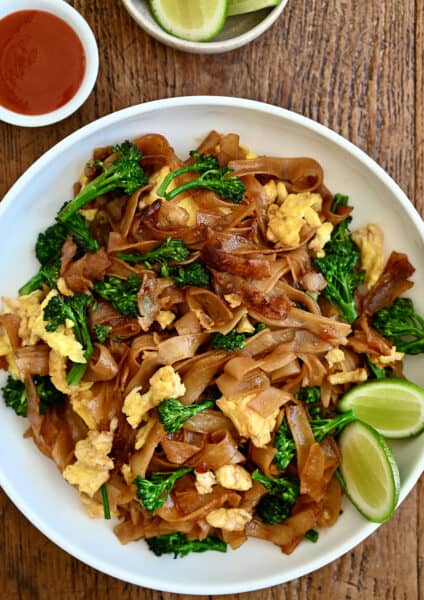 Pad See Ew with lime wedges on a white dinner plate next to a small bowl containing sweet and sour sauce.