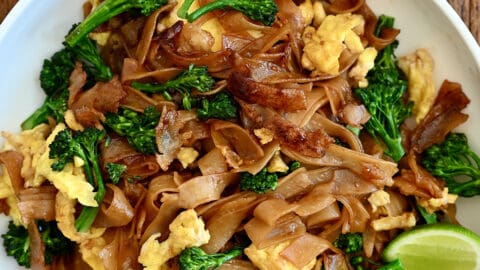 Pad See Ew with lime wedges on a white dinner plate next to a small bowl containing sweet and sour sauce.