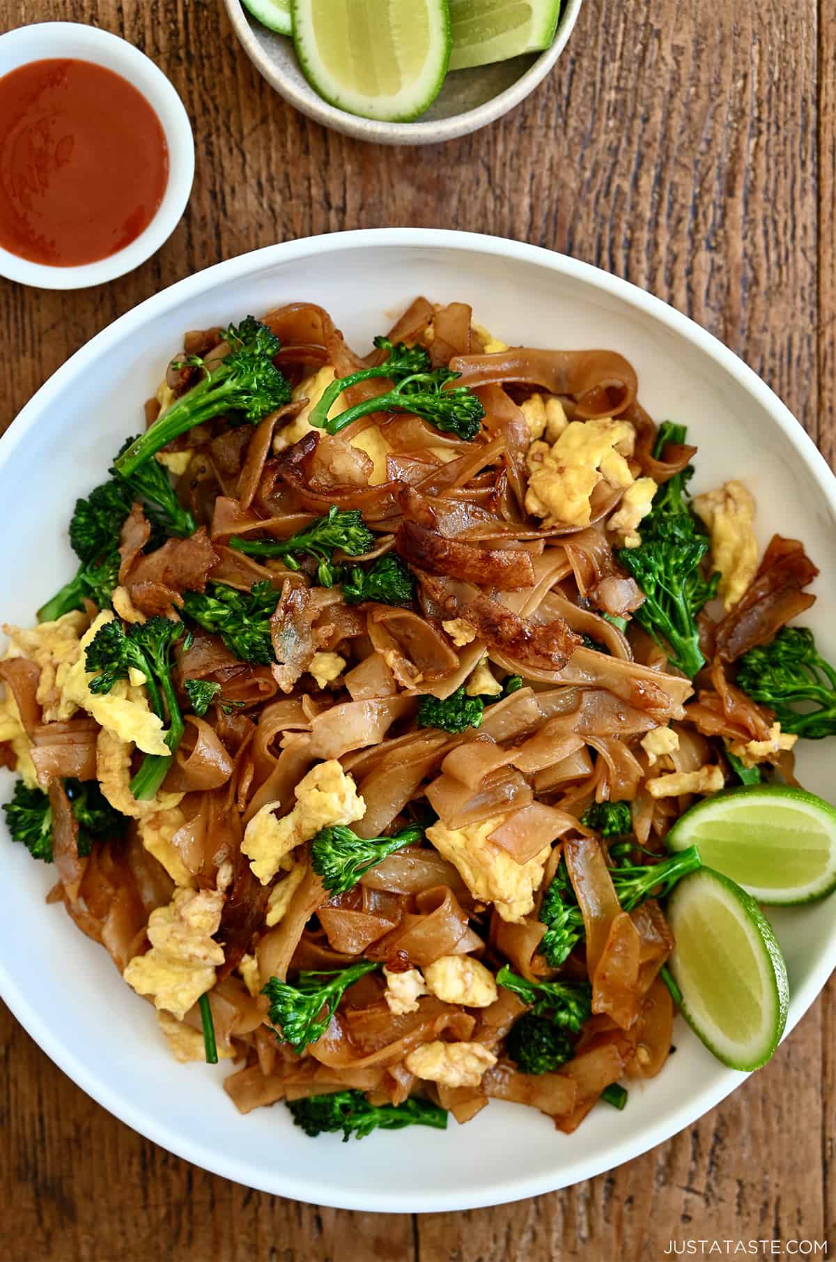 Pad See Ew with lime wedges on a white dinner plate next to a small bowl containing sweet and sour sauce.