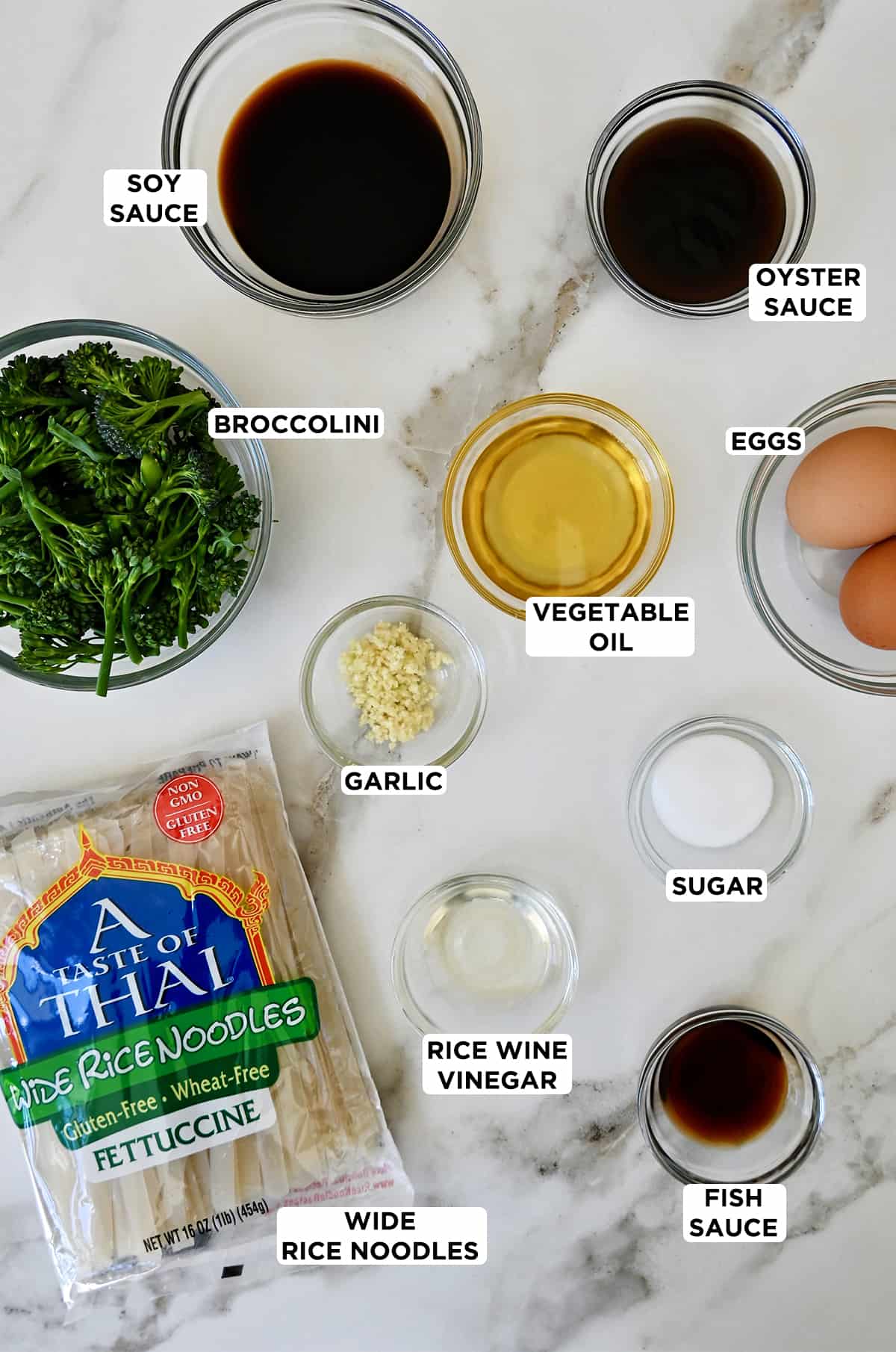 Dried rice noodles next to various sizes of glass bowls containing broccolini, soy sauce, oyster sauce, eggs, vegetable oil, garlic, sugar, rice wine vinegar and fish sauce.