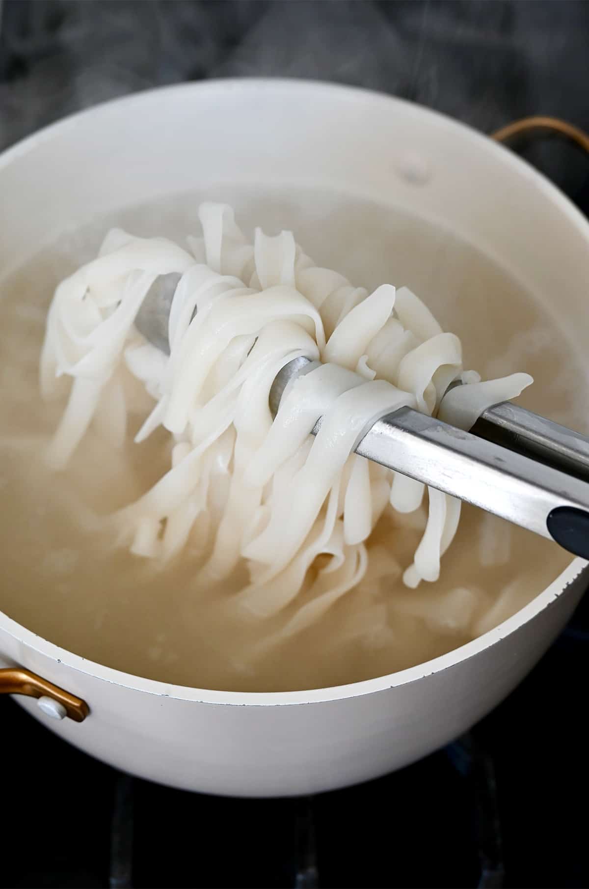 A tongs holds large flat rice noodles out of a boiling pot of water.