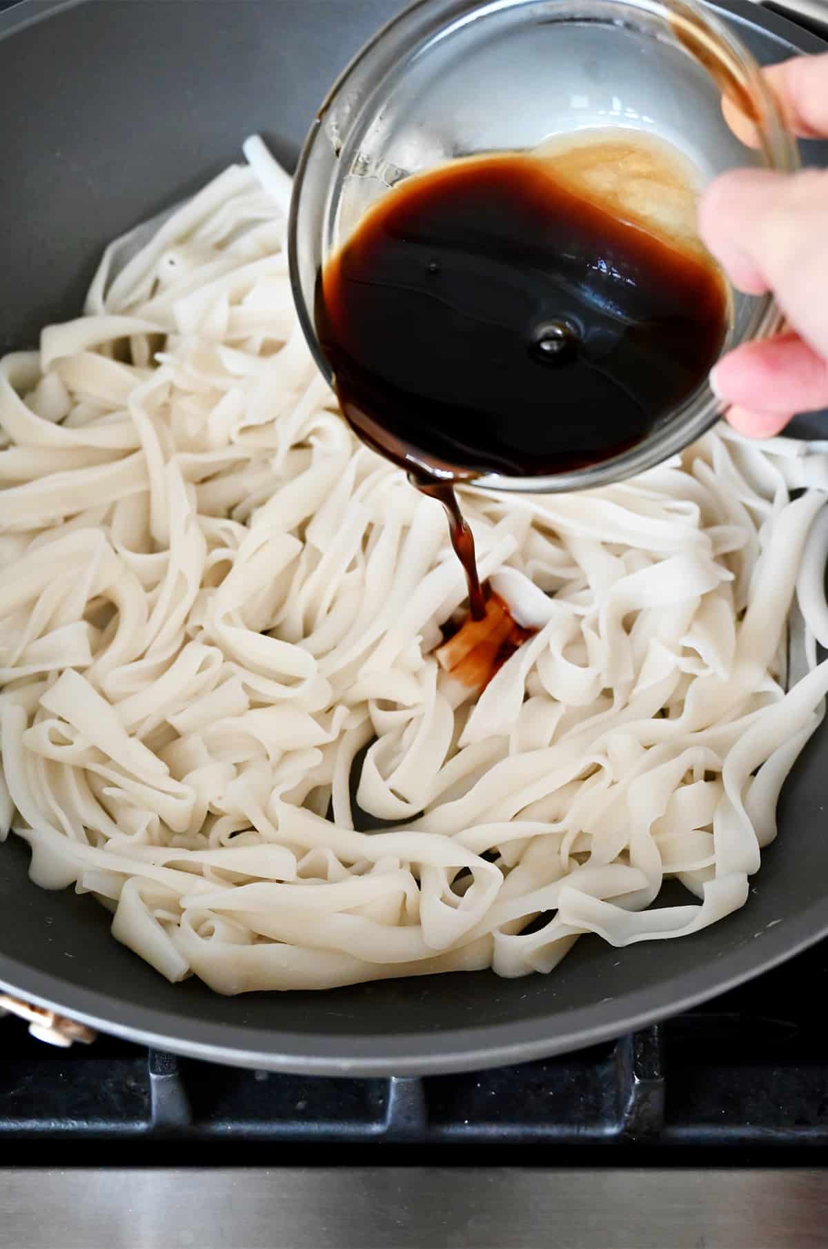 Pad See Ew sauce being poured over cooked rice noodles in a wok.