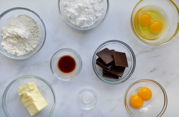 Small bowls containing confectioners' sugar, flour, eggs, chocolate, vanilla extract, salt and butter