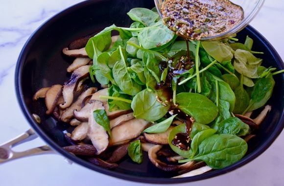 Spicy garlic sauce being poured over shiitakes and fresh spinach in large non-stick skillet