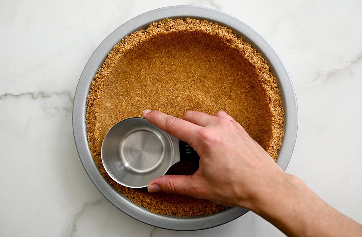 A hand holds a measuring cup atop crushed graham crackers in a pie plate to form a crust