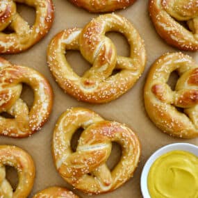 A top-down view of Easy Homemade Soft Pretzels on brown parchment paper next to a small bowl containing yellow mustard