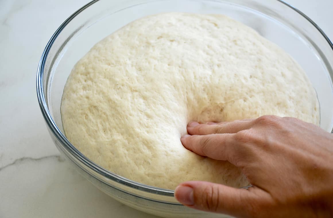 A hand presses into proofed dough in a clear bowl