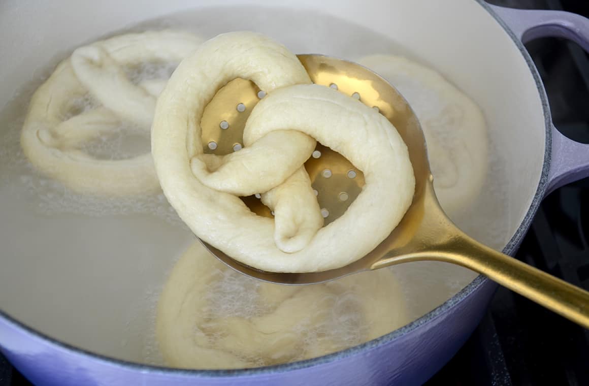 An unbaked pretzel resting atop a slotted spoon above a stockpot filled with a baking soda bath