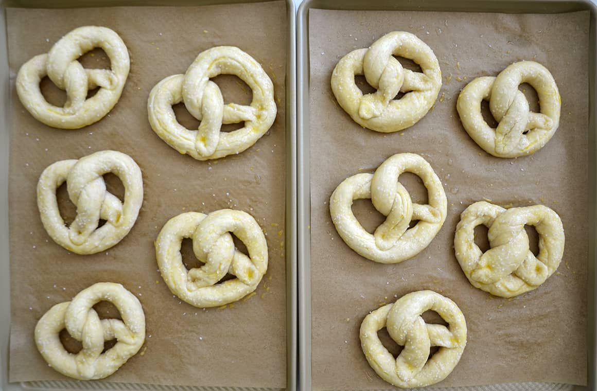A top-down view of unbaked shaped soft pretzels on two parchment paper-lined baking sheets