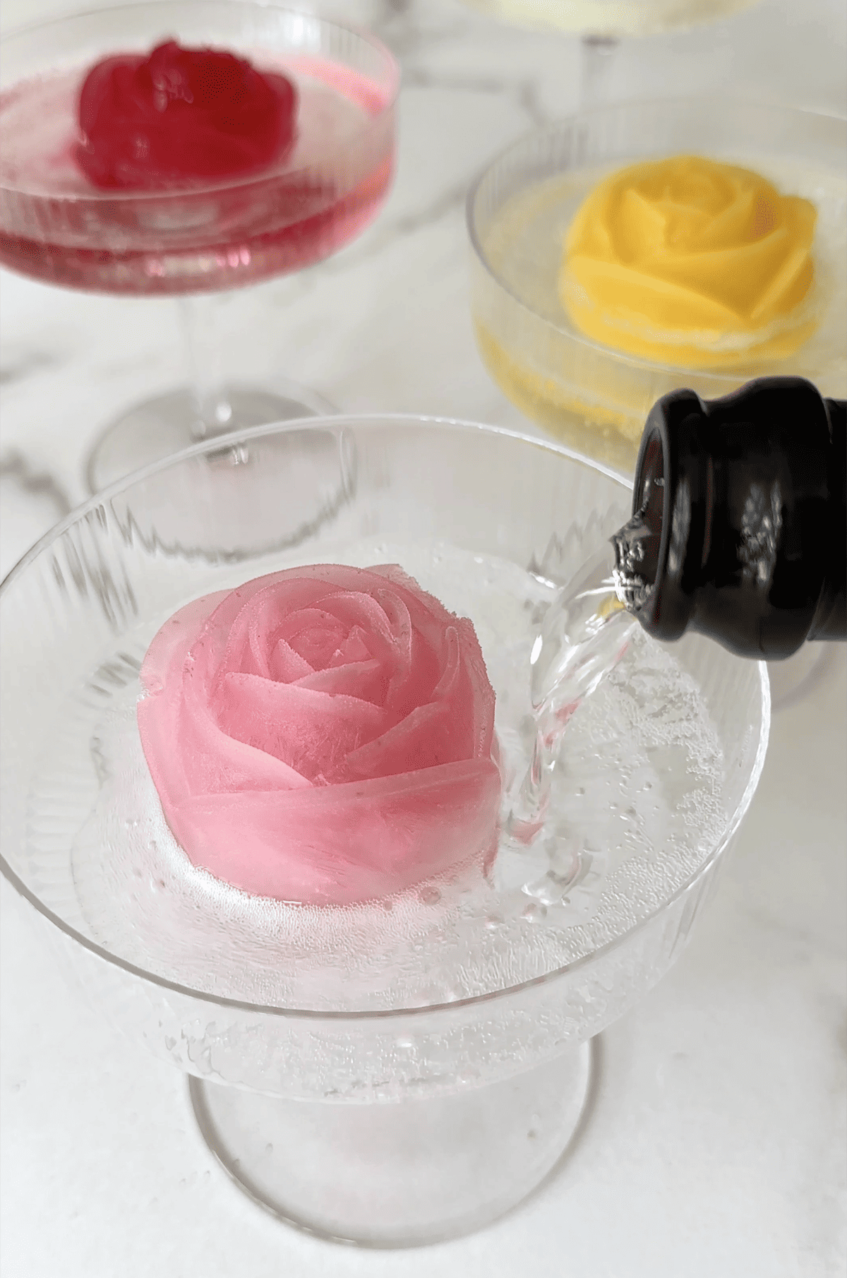 Champagne being poured over a lemonade ice cube in the shape of a rose in a cocktail glass.