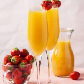 To champagne flutes filled with mimosas garnished with a strawberry next to a bowl containing fresh strawberries.
