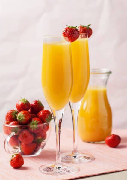 To champagne flutes filled with mimosas garnished with a strawberry next to a bowl containing fresh strawberries.