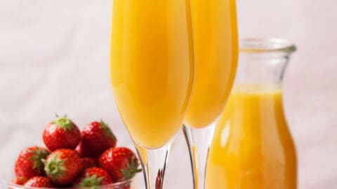 To champagne flutes filled with mimosas garnished with a strawberry next to a bowl containing fresh strawberries.