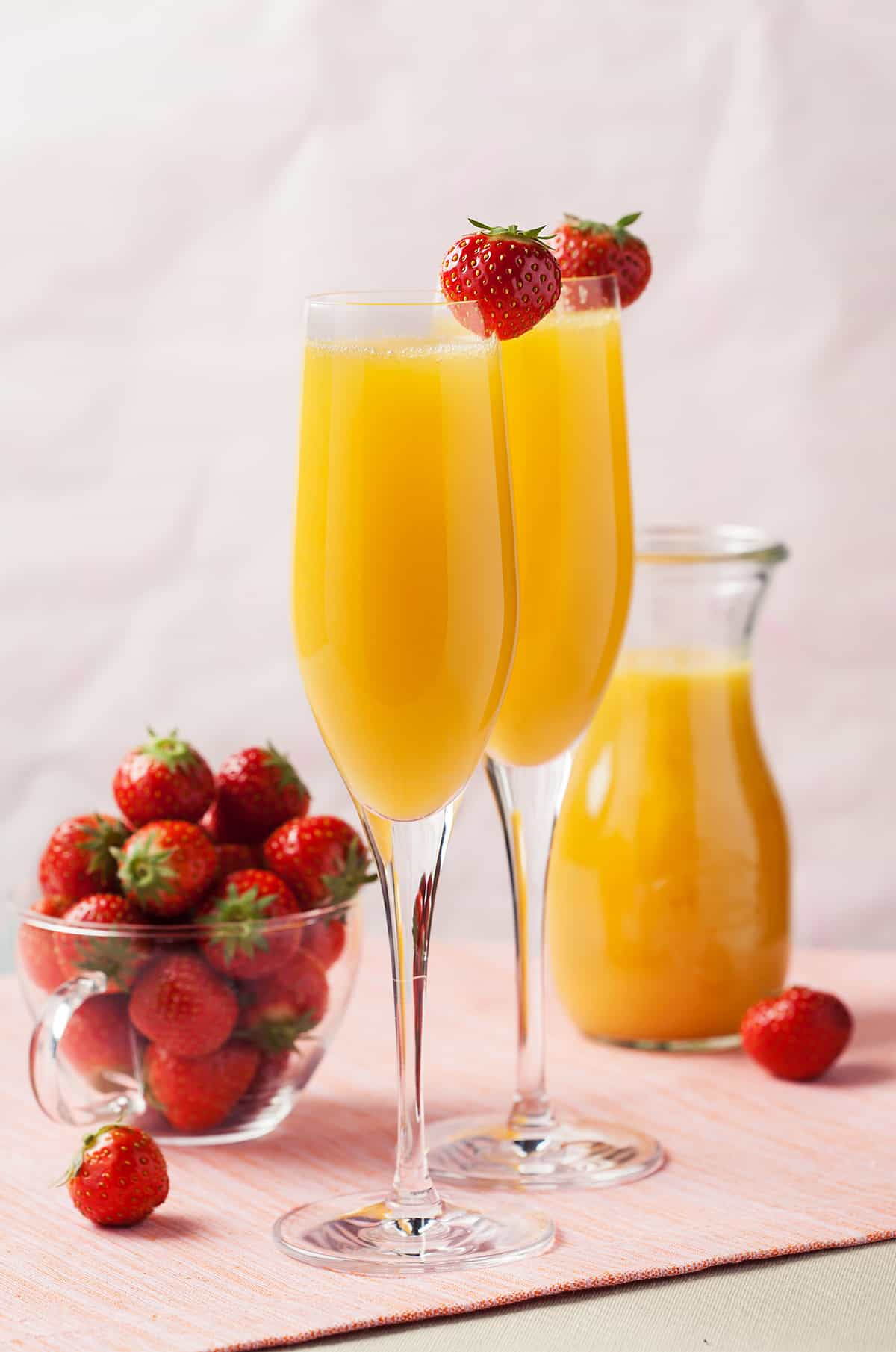 To champagne flutes filled with mimosas garnished with a strawberry next to a bowl containing fresh strawberries.