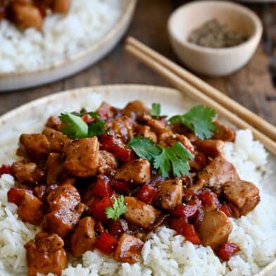 Black pepper chicken over white rice on a plate with chopsticks.