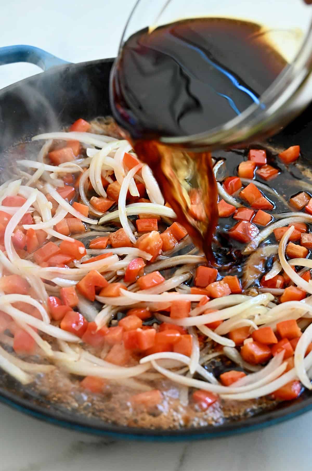 Black pepper sauce being poured into a hot skillet with chopped red bell peppers and onions.
