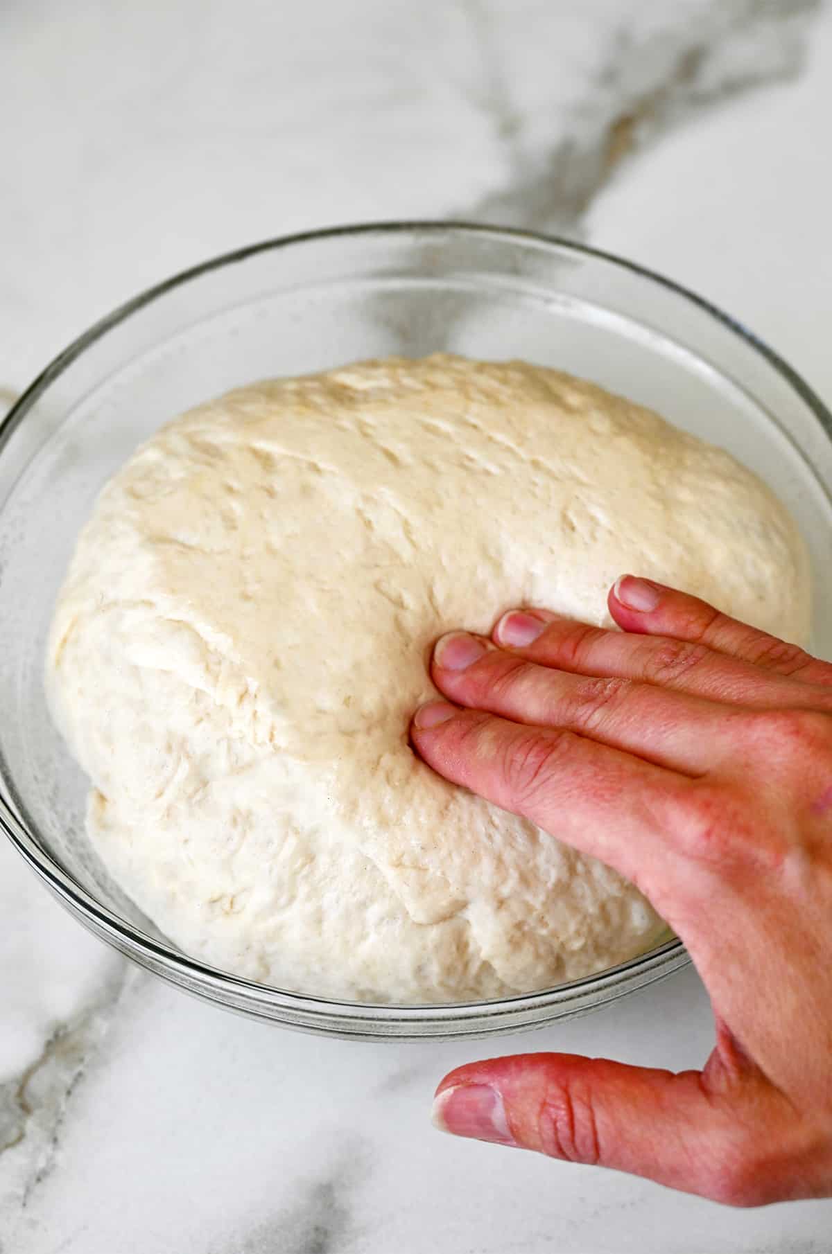 A hand presses into proofed dough in a glass bowl.