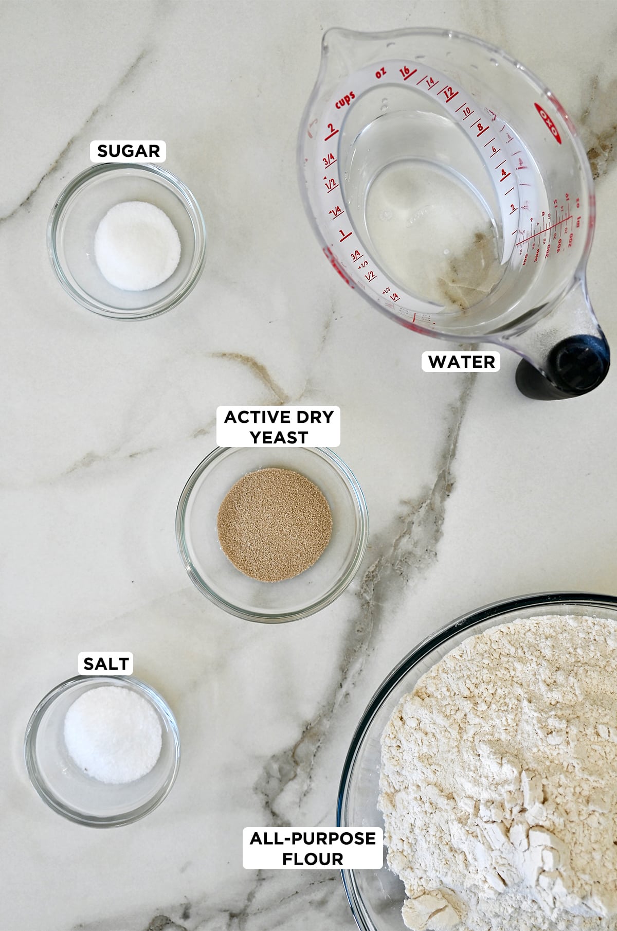 Glass bowls containing sugar, active dry yeast, salt and all-purpose flour next to a liquid measuring cup filled with water.