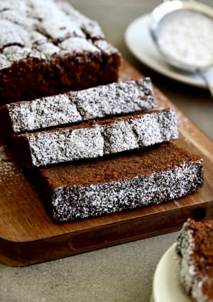 Thick slices of moist chocolate loaf cake with a tight crumb and dusting of powdered sugar on a wooden serving board.