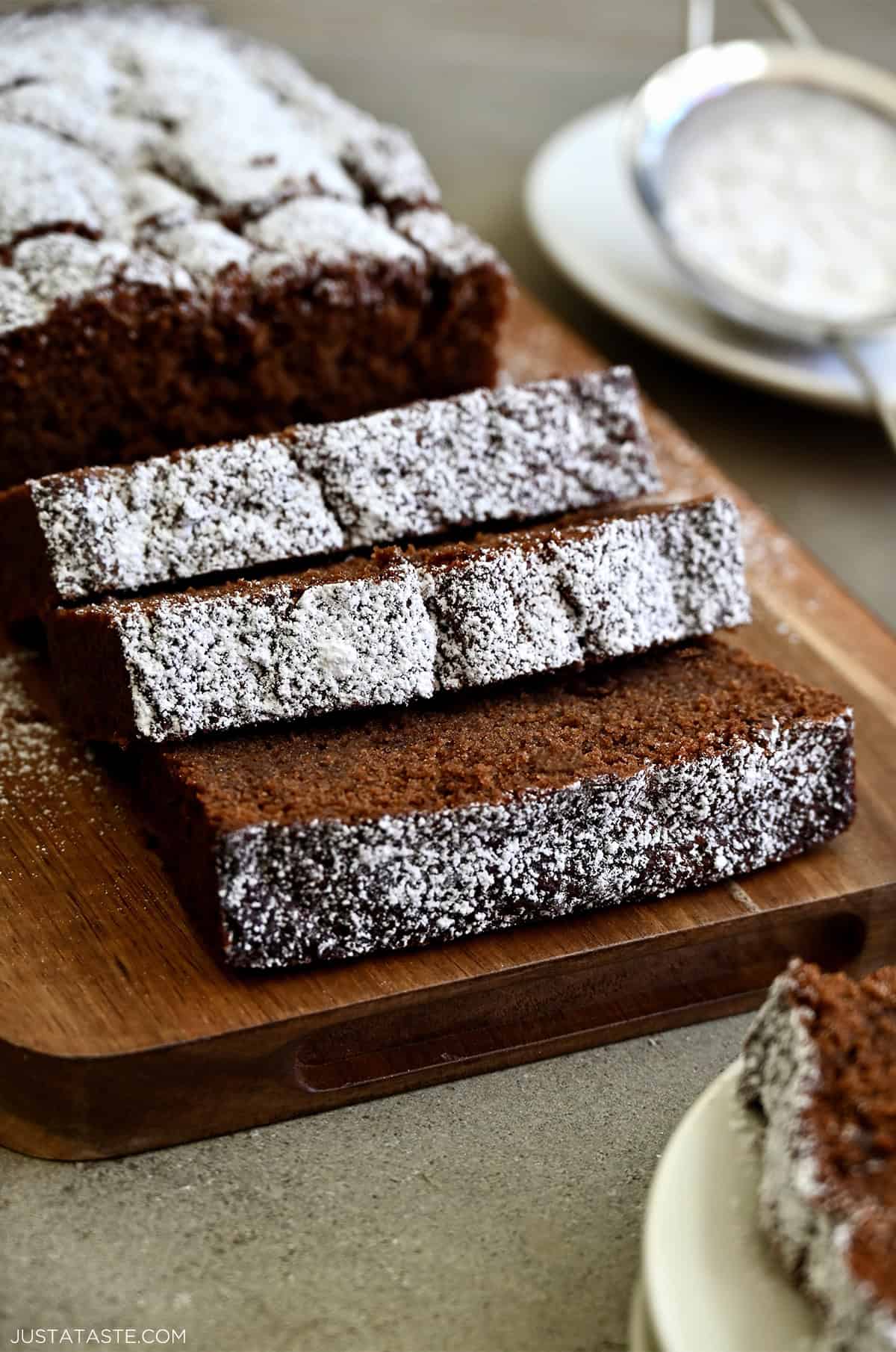 Thick slices of moist chocolate loaf cake with a tight crumb and dusting of powdered sugar on a wooden serving board.