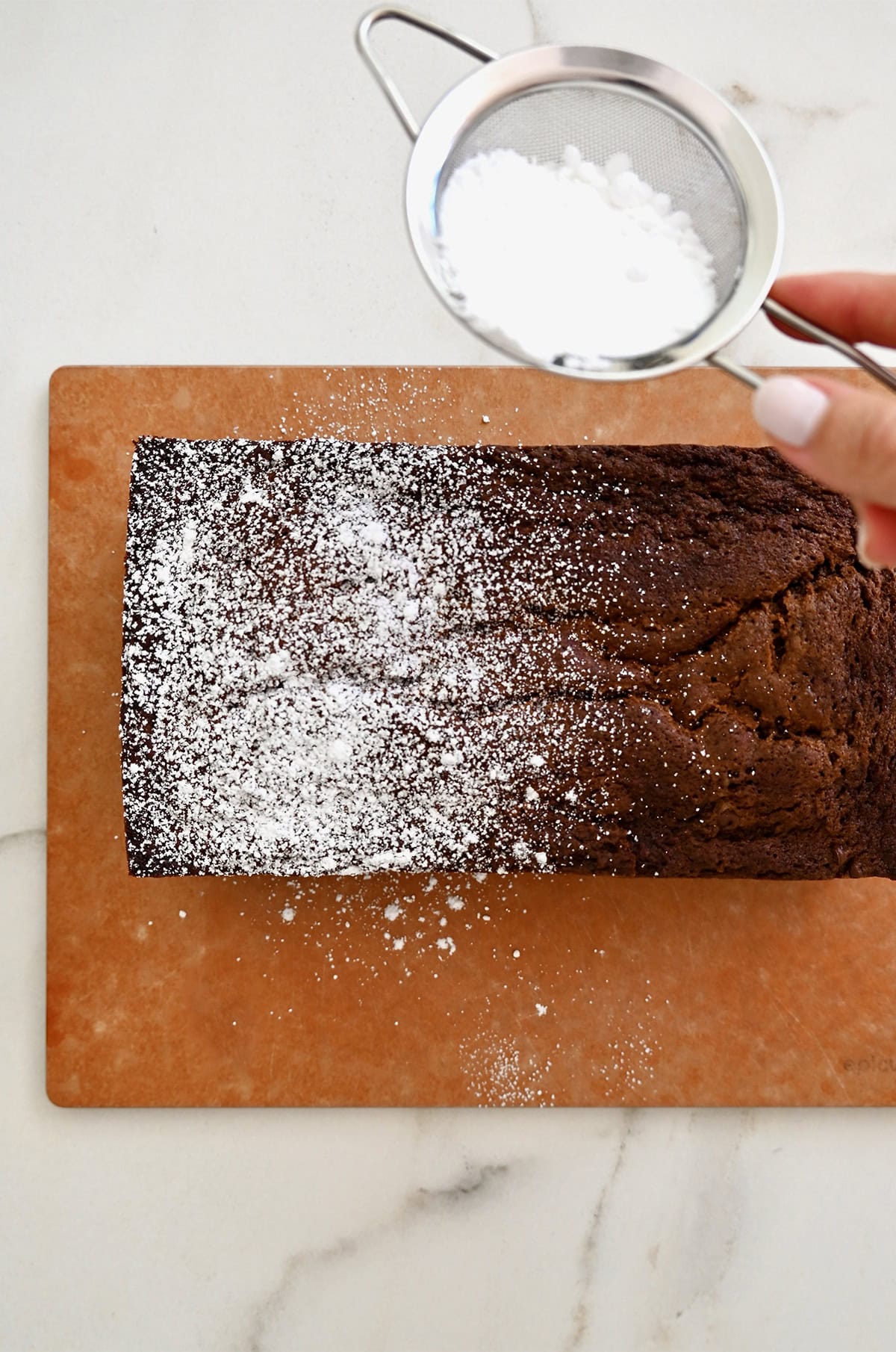 A hand holding a small metal sieve, dusting powdered sugar over the top of a chocolate loaf cake studded with mini chocolate chips.
