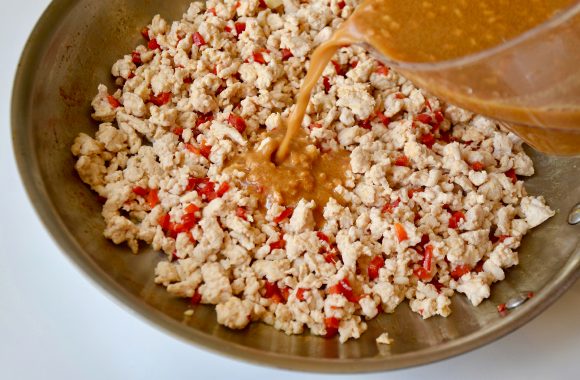 Large sauté pan containing ground turkey and diced red pepper with peanut sauce being added