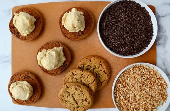 A cutting board containing cookies, ice cream and toppings for ice cream sandwiches