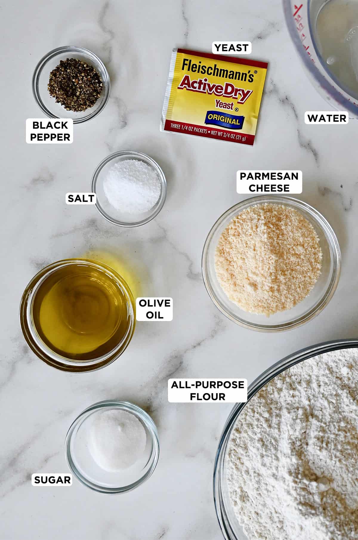 A packet of active dry yeast next to various sizes of glass bowls containing water, Parmesan cheese, flour, sugar, olive oil, salt and black pepper. 
