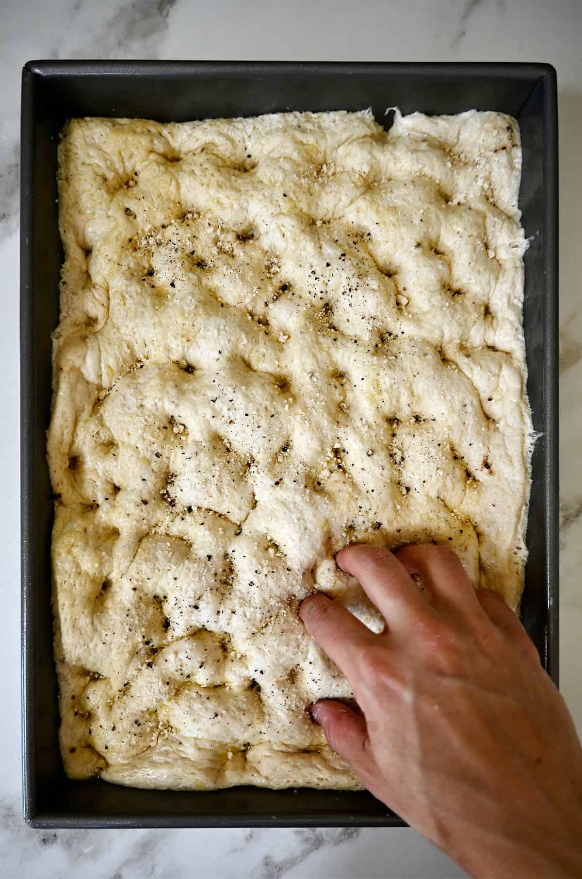 Fingers being pressed into dough in a metal baking pan, creating dimples across the surface. 