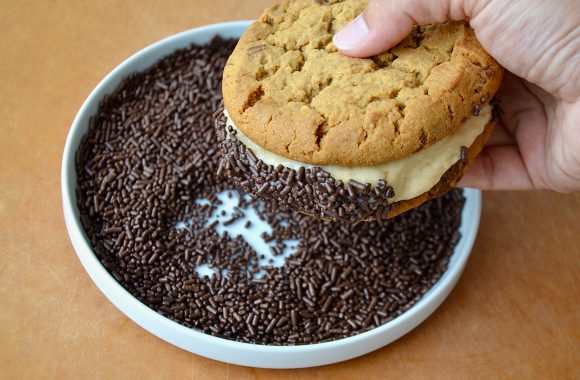 A cookie sandwich being rolled in chocolate sprinkles on a white plate