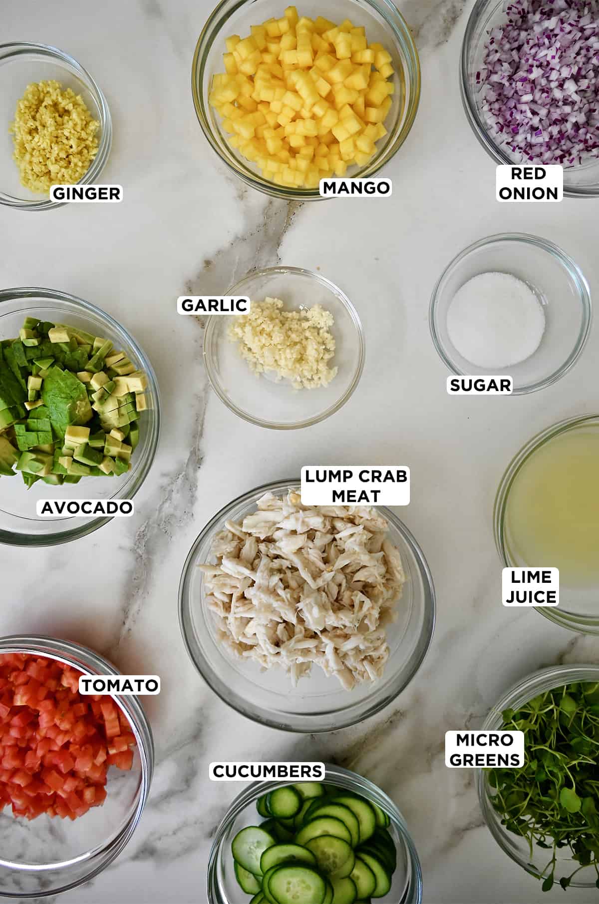 Various sizes of glass bowls containing ingredients to make crab stacks, including diced mango, diced red onion, minced garlic and ginger, sugar, lump crab meat, diced avocado, lime juice, diced tomato and micro greens.