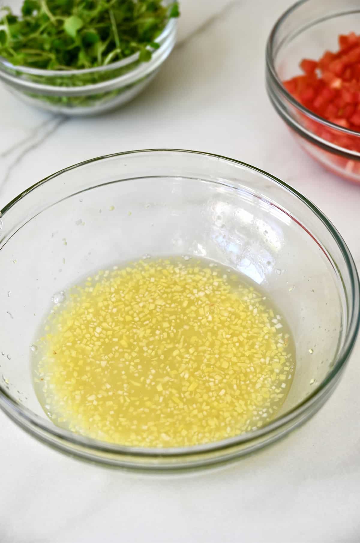 Ginger-lime dressing in a glass bowl next to glass bowls containing micro greens and diced tomatoes.