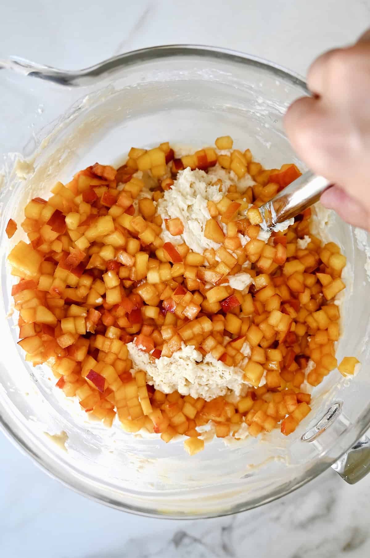 Diced peaches being folded into cupcake batter in a large glass bowl.